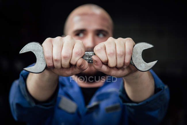 Mecánico masculino sin rostro en la cara de recubrimiento general azul con llave de metal en las manos mirando hacia otro lado sobre fondo negro - foto de stock