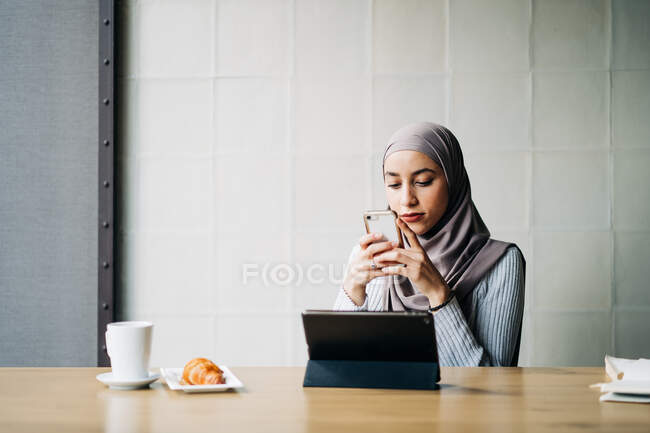 Vista laterale della freelance musulmana che indossa hijab tradizionale in piedi nel caffè e naviga smartphone mentre lavora al progetto in remoto — Foto stock