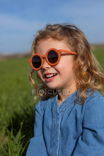 Мила щаслива дівчинка в модному одязі і сонцезахисних окулярах сидить і розслабляється на трав'янистому газоні — стокове фото