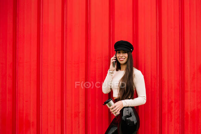 Serious stylish female entrepreneur leaning on red wall in city and discussing project on smartphone while standing with drink to go looking at camera — Stock Photo