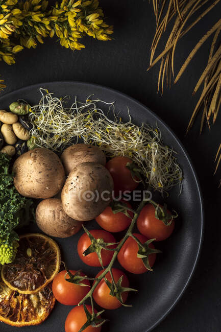 Vue du dessus de l'assiette avec paquet de tomates cerises fraîches près de pommes de terre cuites et germes sur fond gris avec des fleurs — Photo de stock