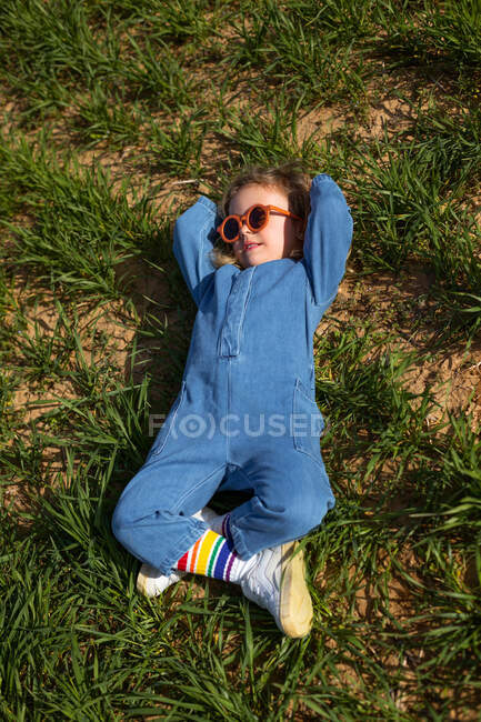 Dall'alto ragazza in abiti alla moda e occhiali da sole che si tiene per mano dietro la testa e rilassarsi sul prato erboso — Foto stock