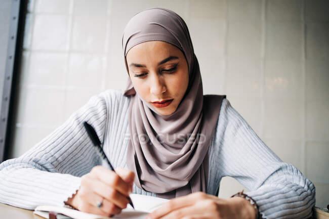 Сконцентрована мусульманка-фрилансер в традиційному головному шарфі сидить за столом і пише в блокноті під час роботи в кафе. — стокове фото