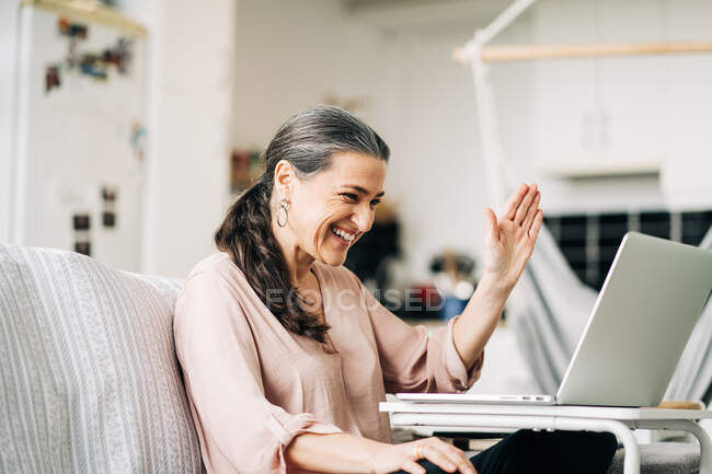 Positive middle aged female sitting on couch waving while having video chat on netbook in modern apartment — Stock Photo