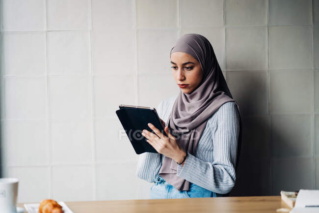 Vista lateral do freelancer muçulmano vestindo hijab tradicional em pé no café e tablet de navegação enquanto trabalhava no projeto remotamente — Fotografia de Stock