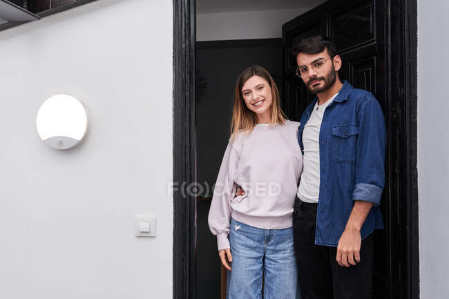 Feliz joven pareja multiétnica en ropa casual acurrucándose y mirando a la cámara mientras está de pie en la puerta del apartamento moderno - foto de stock