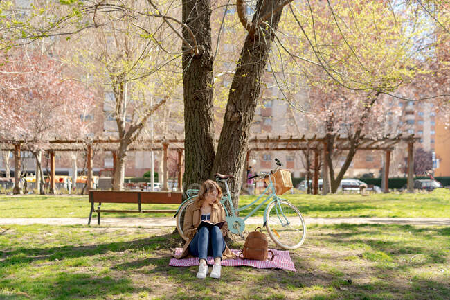 Corpo pieno di concentrato giovane femmina prendere appunti nel quaderno su tessuto a scacchi con zaino vicino bicicletta nel parco — Foto stock