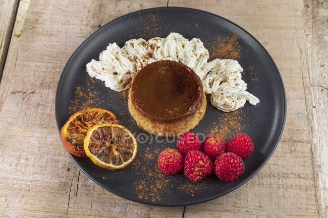 De arriba del flan sabroso con la crema batida y los trozos rojos anaranjados con la canela en polvo sobre el plato cerámico - foto de stock