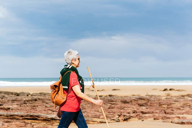 Side view of elderly female backpacker with trekking pole strolling on boulders against stormy ocean under cloudy sky — Stock Photo