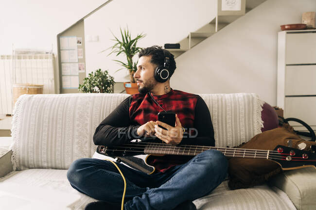 Erwachsene bärtige männliche Musiker mit Bassgitarre in Kopfhörer SMS auf Handy im Wohnzimmer — Stockfoto