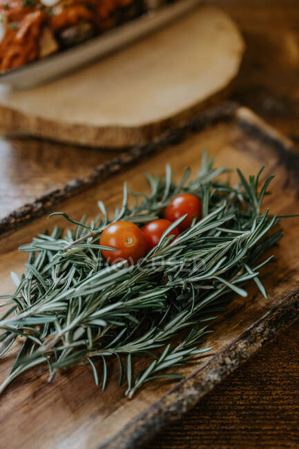 De arriba de los tomates enteros maduros de cereza sobre las ramitas frescas de romero con el olor agradable en el plato de madera - foto de stock