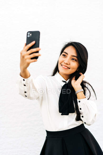 Femme asiatique avec de longs cheveux noirs prenant autoportrait sur téléphone portable debout sur fond blanc — Photo de stock