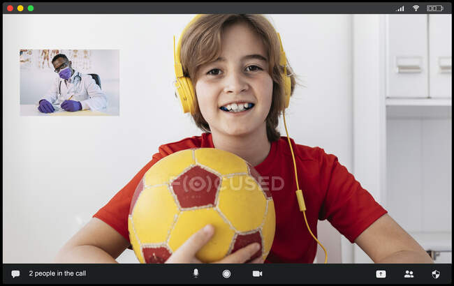 Delighted boy in headphones with ball in hand smiling while having video chat with unrecognizable African American male doctor in medical robe and mask taking notes during distance consultation — Stock Photo