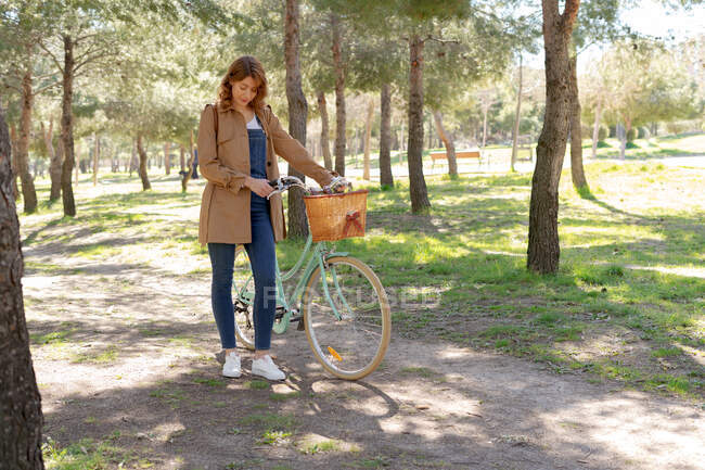 Corpo cheio de jovem fêmea em pé e concentrado perto de bicicleta velha com cesta de vime de madeira no parque — Fotografia de Stock