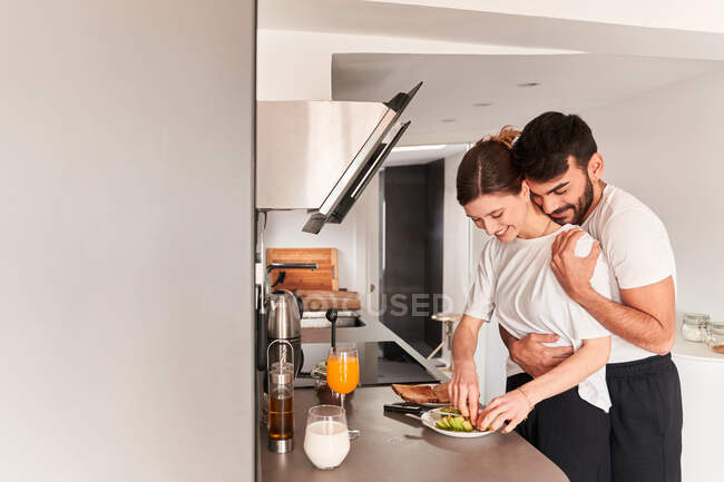 Vista laterale del giovane ragazzo etnico romantico in t shirt bianca sorridente e abbracciante fidanzata felice preparare una colazione sana con avocado in cucina — Foto stock