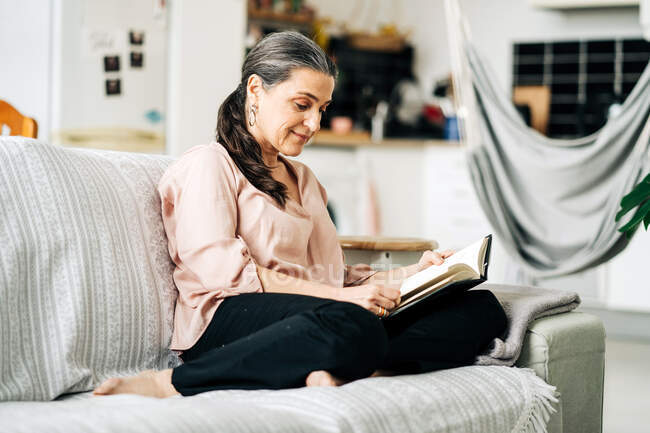 Повне тіло босоногих жінок читає книгу, сидячи на зручному дивані у вітальні з зеленою рослиною вдома — стокове фото