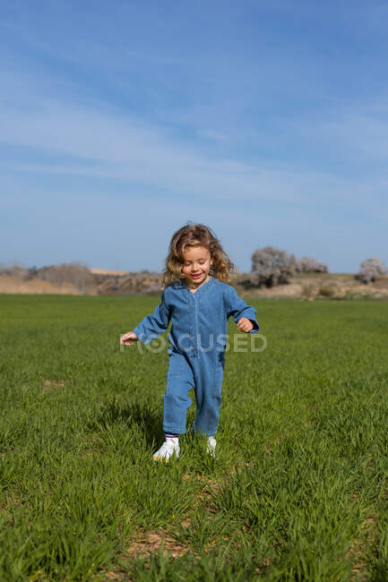 Fille ravie avec les cheveux bouclés souriant et courant sur l'herbe verte contre le ciel bleu le jour d'été dans le champ — Photo de stock