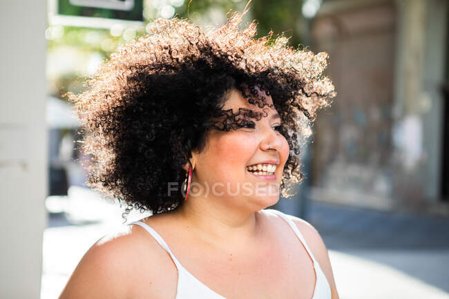 Sonriente adulto regordeta hembra con Afro peinado mirando hacia otro lado en la ciudad en la espalda iluminado - foto de stock