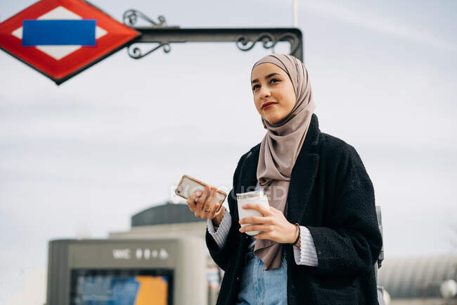 Приємна етнічна жінка в хіджабі і модний одяг стоять з випивкою під час випивки в інтернеті на смартфоні і насолоджуються вихідними в місті. — стокове фото