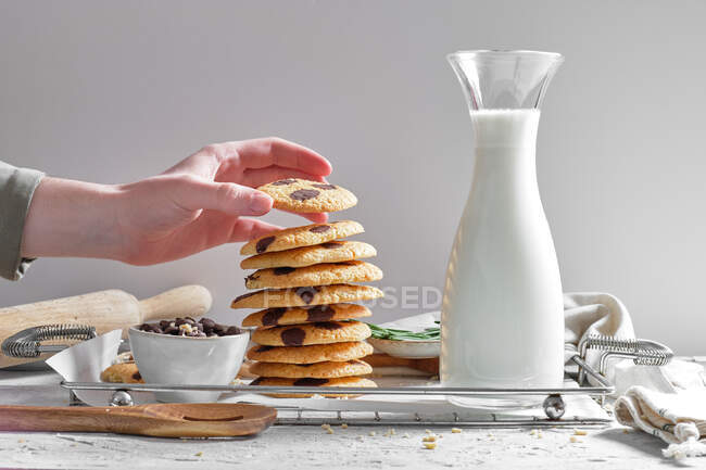 Crop anonymous housewife stacking delicious homemade cookies with chocolate chips on tray with jar of fresh milk in kitchen — Stock Photo