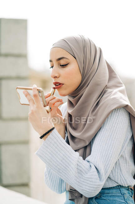 Vue latérale de la femme musulmane en foulard appliquant du rouge à lèvres et regardant l'écran du smartphone tout en faisant du maquillage dans la rue — Photo de stock