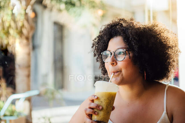 Mujer alegre adulta en gafas sentada en la mesa de la cafetería urbana con un vaso de bebida mientras mira hacia otro lado - foto de stock