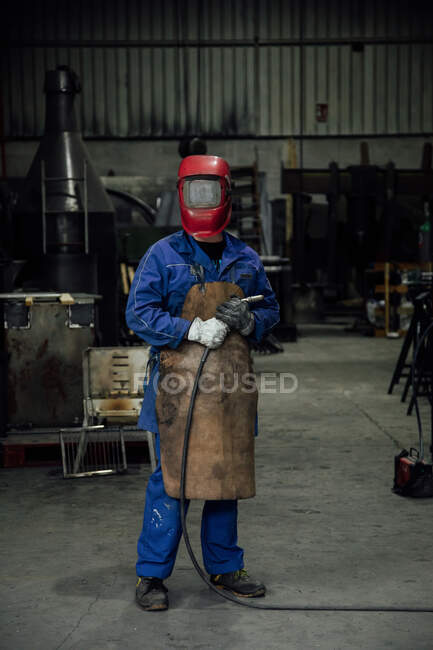 Mecánico anónimo en casco protector y delantal en azul de pie general con manguera de soldadura en taller de luz cerca de construcciones metálicas - foto de stock