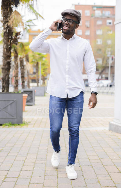 Maschio afroamericano alla moda che cammina per strada con palme e telefono cellulare parlante — Foto stock