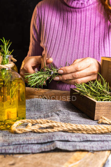 Crop unrecognizable woman in purple sweater cutting herbs twigs with scissors at table with essential oil glass bottles and rope with small chest on textile — Stock Photo
