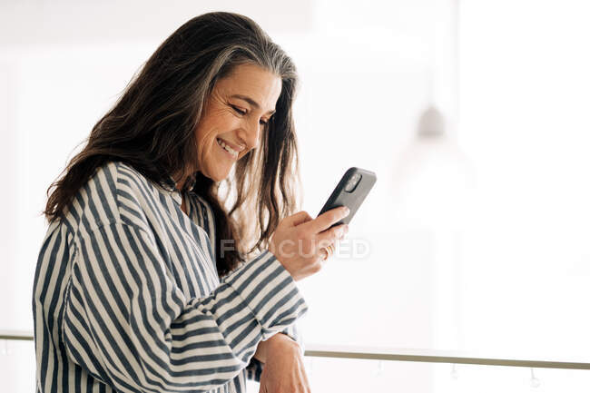 Seitenansicht positiver weiblicher Textnachrichten mittleren Alters auf dem Handy, während sie im Raum mit kreativer Malerei und Staffelei auf verschwommenem Hintergrund sitzt — Stockfoto
