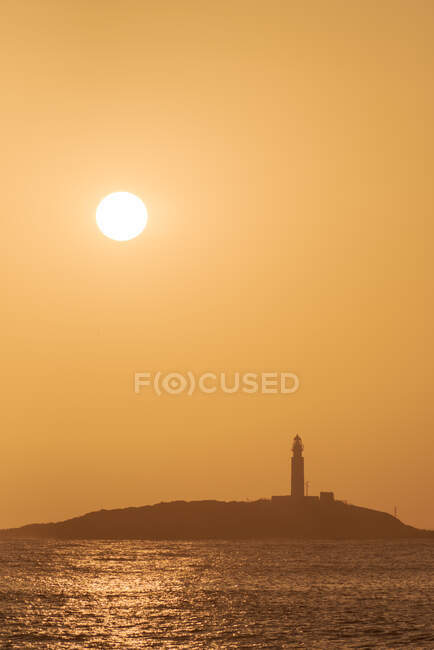 Pintoresco paisaje de aguas onduladas de la costa de lavado oceánico con faro de gran altura situado en Faro de Trafalgar en Cádiz en España bajo un cielo naranja brillante al amanecer - foto de stock