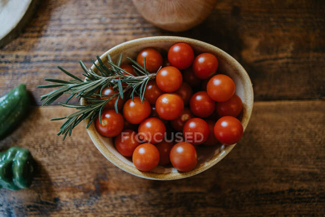 Schüssel mit frischen Kirschtomaten in der Nähe von Rosmarinstielen und ganzen Zwiebeln auf Holztisch — Stockfoto