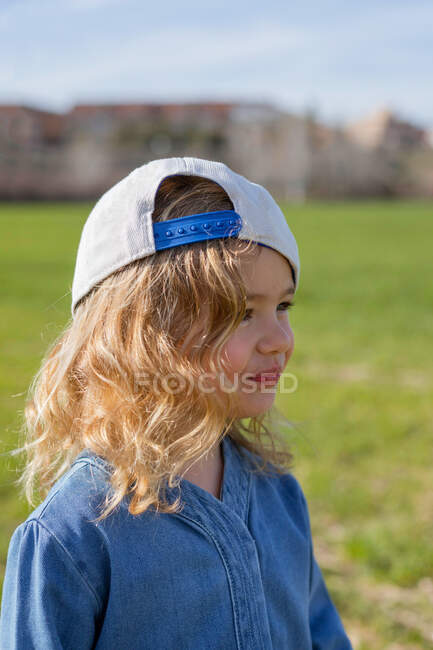 Side view of cute girl in stylish clothes and cap looking away while standing on grass on sunny summer day in field — Stock Photo
