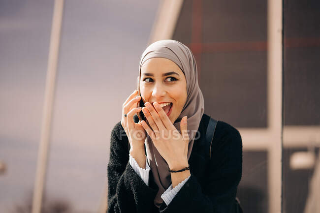 Femme ethnique positive dans le hijab debout dans la rue de la ville et avoir une conversation sur un téléphone portable tout en détournant les yeux — Photo de stock