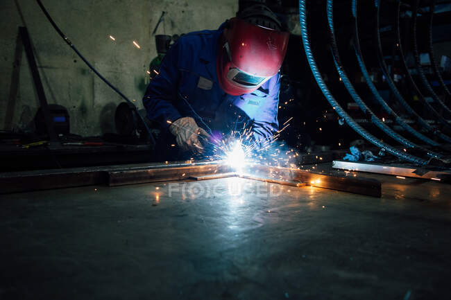 Crop faceless worker in gloves and uniform welding metal details on table near constructions in factory — Stock Photo