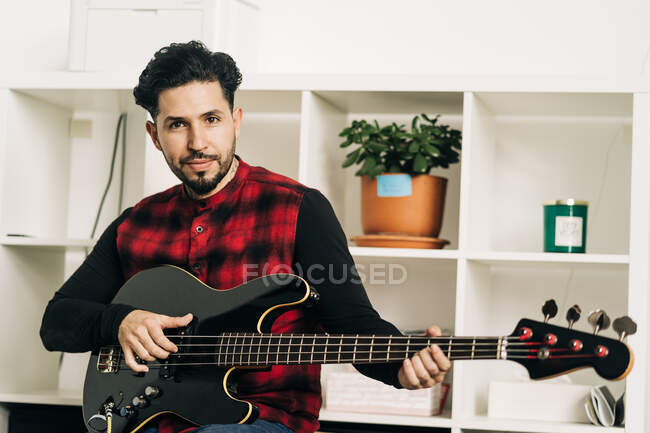Adult professional bearded male musician playing bass guitar during rehearsal against shelves at home — Stock Photo