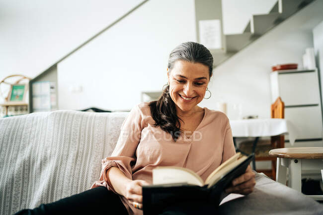 Livro de leitura feminina feliz enquanto sentado em sofá confortável na sala de estar com planta verde em casa — Fotografia de Stock