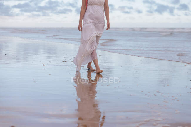 Vue latérale d'une femelle anonyme se promenant dans l'eau ondulée d'un vaste océan sur une plage de sable fin sous un ciel nuageux — Photo de stock