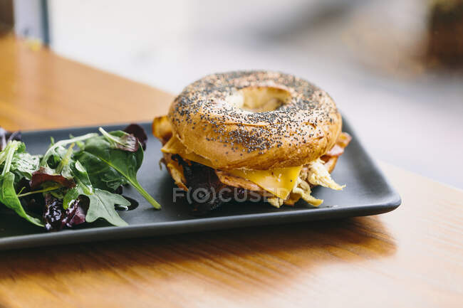 Sandwich bagel appétissant au fromage et poulet servi dans une assiette avec salade de rucola sur la table dans un café — Photo de stock