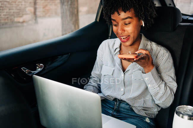 Jovem afro-americana ocupada gravando mensagem de voz no celular e trabalhando com netbook no automóvel no banco de trás — Fotografia de Stock