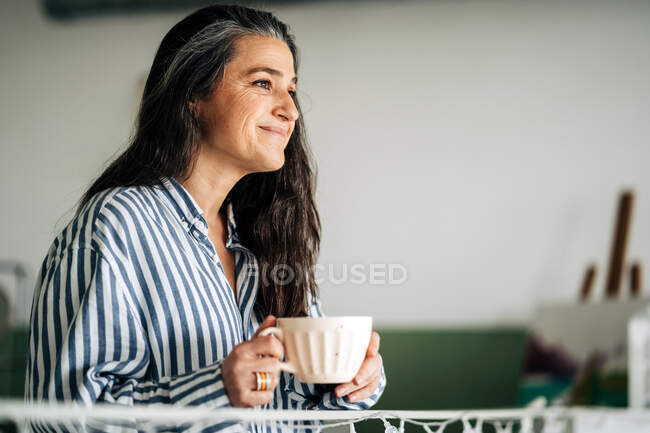 Side view of smiling middle aged female with cup of hot beverage looking into distance while sitting in room with paintings and easel on blurred background — Stock Photo