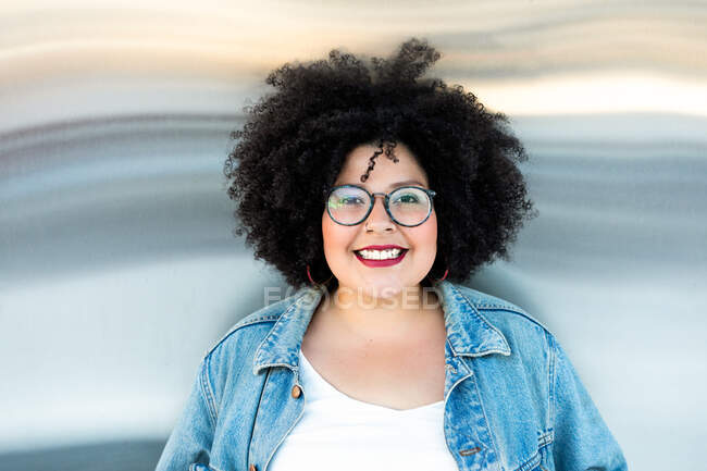 Adult overweight female in trendy clothes and eyewear with Afro hairstyle looking at camera on blurred background — Stock Photo