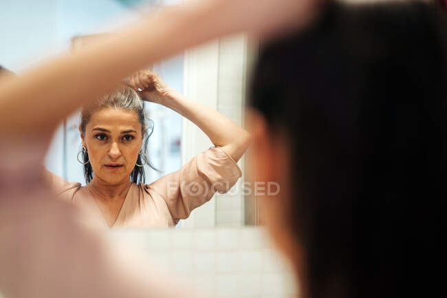 Сосредоточенная женщина средних лет смотрит на отражение в зеркале, делая конский хвост в светлом помещении дома во время повседневной рутины. — стоковое фото
