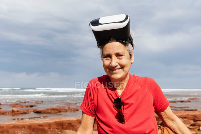 Content old female tourist in virtual reality headset looking at camera on ocean coast under cloudy sky — Stock Photo