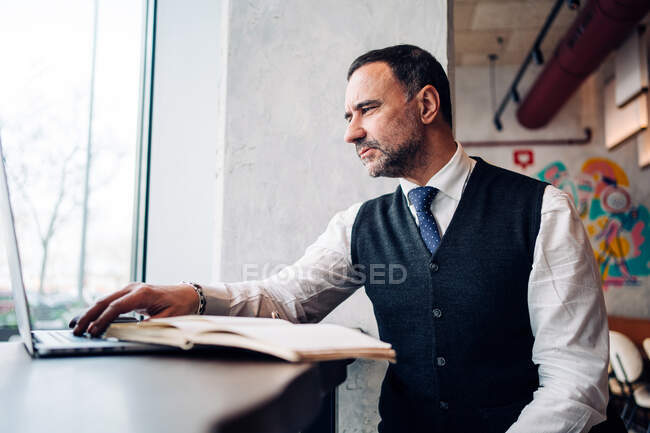 Maturo ispanico maschio imprenditore in abiti formali lavorando su netbook al tavolo del caffè con giornale — Foto stock