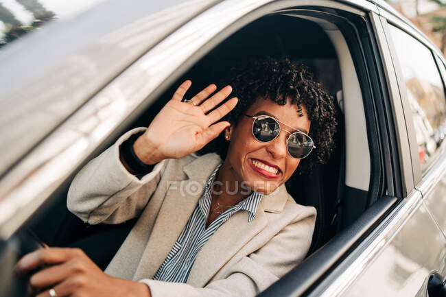 Cheerful African American female in stylish sunglasses and fashionable outfit smiling and waving hand in automobile — Stock Photo