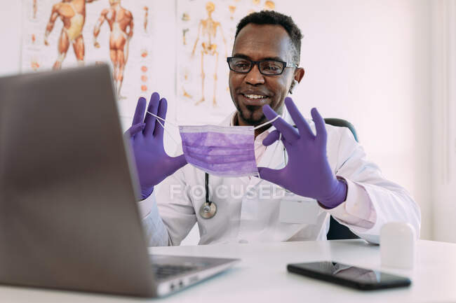Médico masculino afroamericano adulto positivo en uniforme médico y guantes de látex sonriendo y mostrando máscara mientras tiene videoconferencia a través de computadora portátil en el hospital moderno - foto de stock