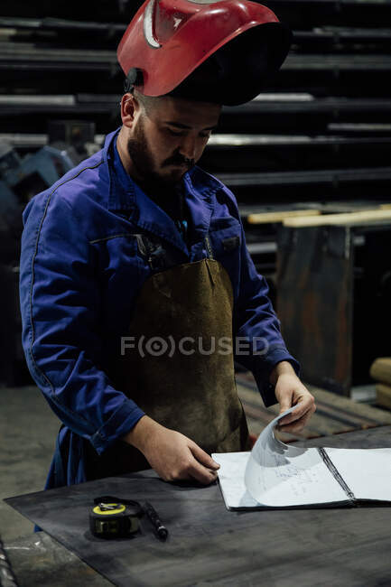 Trabajador masculino barbudo serio en hardhat y buscando notas en cuaderno con pluma en la mesa cerca de construcciones metálicas en garaje ligero - foto de stock