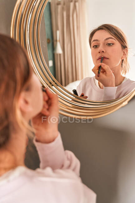 Schnurgerade junge Frau mit blonden Haaren in lässiger Kleidung, die in den Spiegel schaut und Lippenstifte am sonnigen Morgen aufträgt — Stockfoto