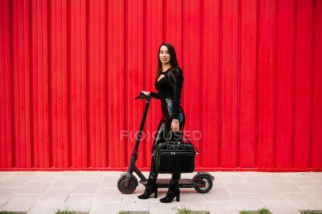 Seitenansicht einer stilvollen Unternehmerin, die mit einem Elektroroller auf dem Bürgersteig in der Stadt gegen eine rote Wand läuft und in die Kamera blickt — Stockfoto